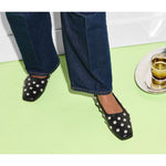 A person wearing jeans and stylish black Freda Salvador Jessie Ballet Flats with silver studs stands on a light green floor. To the right, there's a tray with a glass of amber liquid, completing the scene with a touch of handcrafted elegance.