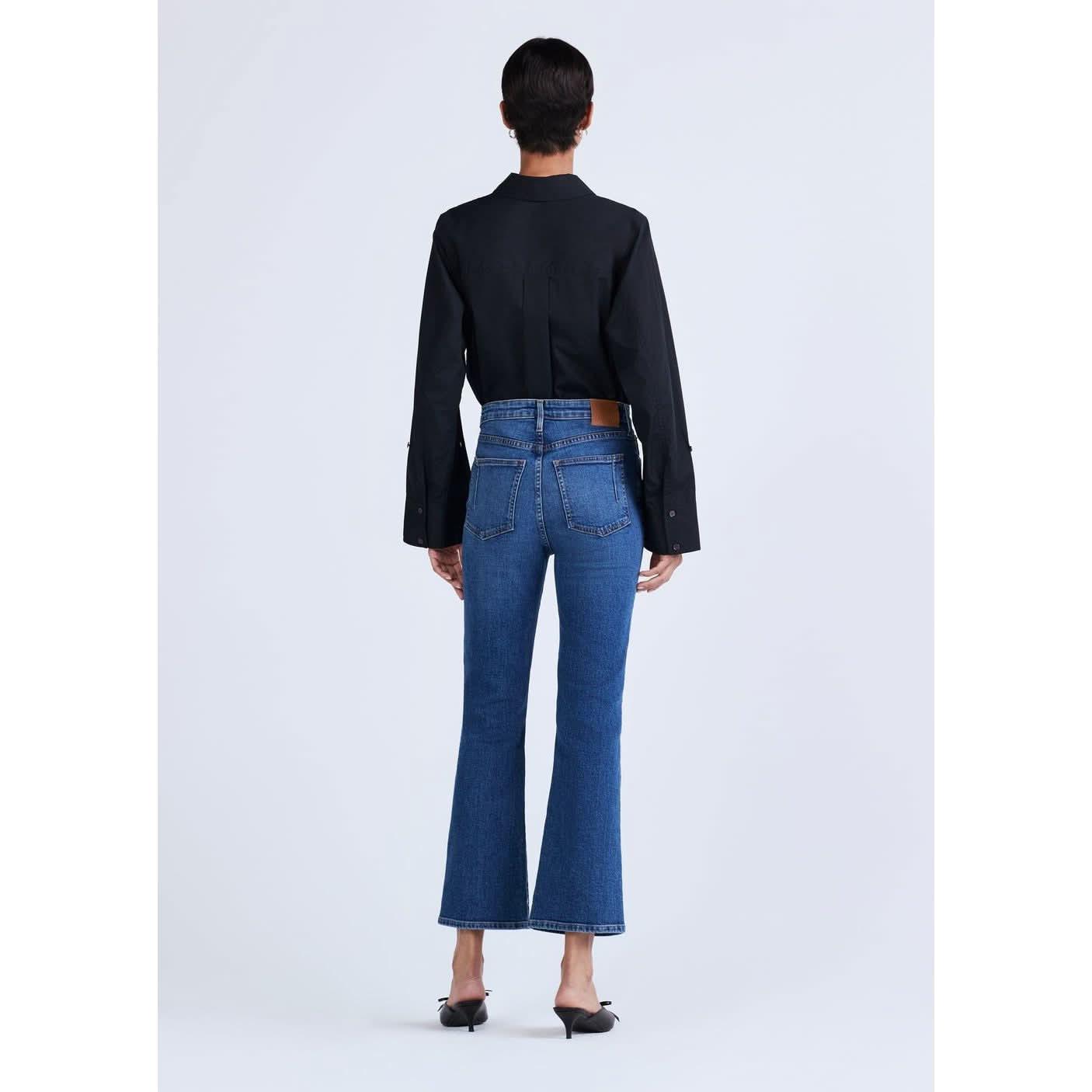 A person with short dark hair is standing with their back to the camera, wearing a black long-sleeved shirt, Derek Lam 10 Crosby High Rise Crop Flare Jeans by Derek Lam in premium denim blue, and black pointed-toe heels. The background is plain white.