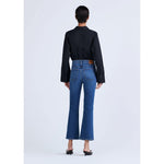 A person with short dark hair is standing with their back to the camera, wearing a black long-sleeved shirt, Derek Lam 10 Crosby High Rise Crop Flare Jeans by Derek Lam in premium denim blue, and black pointed-toe heels. The background is plain white.