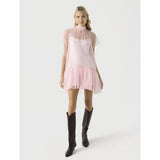 A person is dressed in a Simkhai Beaux Tiered Mini Dress, showcasing a short, flowing pink style with sheer sleeves and a ruffled hem. Their long hair complements the knee-high black boots as they stand against a plain light background.