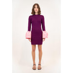 A person stands in a Simkhai Agathea dress, a purple outfit with large pink cuffs. The elegant drape of the knee-length design is complemented by its long sleeves. They wear black, open-toed heels, all set against a plain white background.