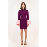 A person stands in a Simkhai Agathea dress, a purple outfit with large pink cuffs. The elegant drape of the knee-length design is complemented by its long sleeves. They wear black, open-toed heels, all set against a plain white background.