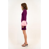 A woman stands in profile, wearing a Simkhai Agathea gown in a celestial shade of purple with oversized pink cuffs. She also dons black heeled sandals, exuding an elegant drape against the plain white background.